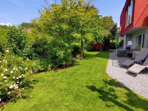 Grüner Garten mit Rasen, Sträuchern und einer Terrasse neben einem roten Haus.
