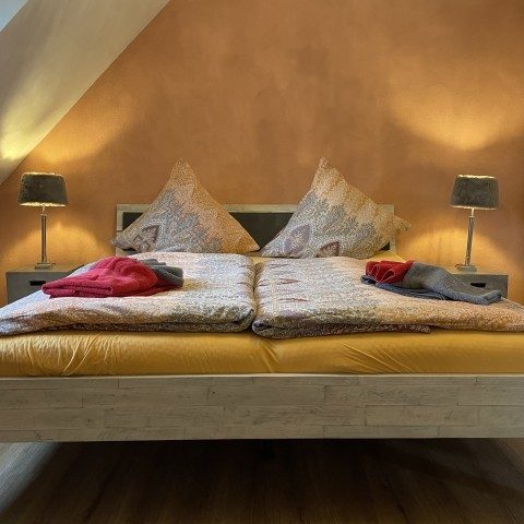 Doppelbett mit Kissen, Decken und Nachttischen in einem warmen, orangefarbenen Raum.