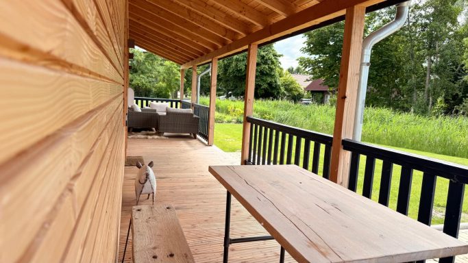 Holzveranda mit Tisch, Blick auf eine grüne Wiese und Sitzmöbel im Hintergrund.