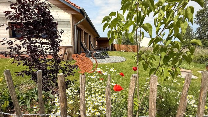 Garten mit Blumen und Hecke, im Hintergrund ein Wohnhaus mit Terrasse.