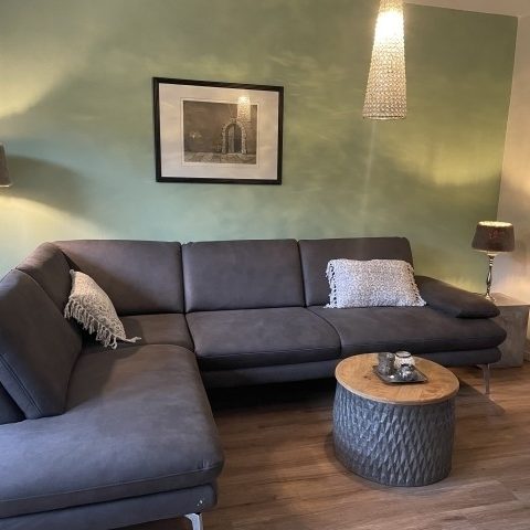Geräumiges Wohnzimmer mit grauem Sofa, Holztisch und stilvollen Lampen an grüner Wand.