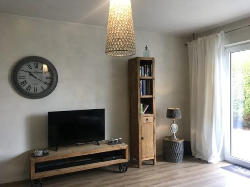 Wohnzimmer mit Fernseher, Regal, Uhr und Lampe, hell mit Fenster und Vorhängen.