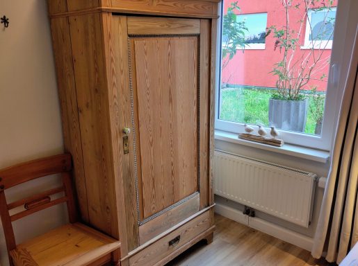Holzschrank mit einer Tür, neben einem Fenster mit Pflanzen und einem Stuhl.